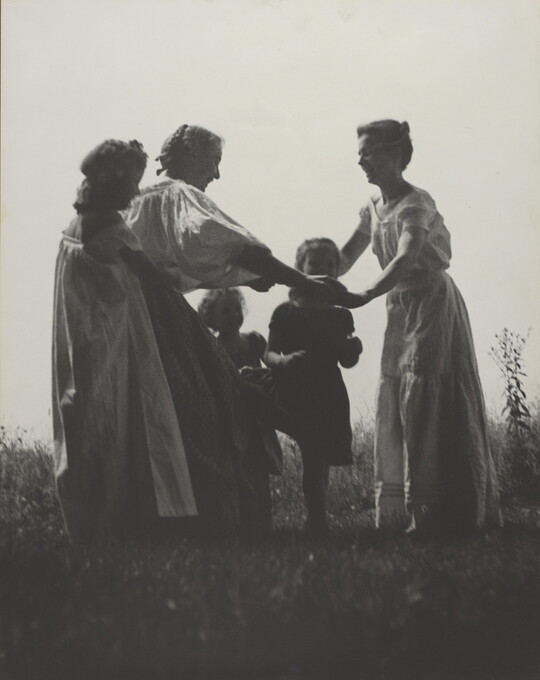 A black-and-white photograph of White women and children wearing long dresses holding hands to form a circle in a grassy field.