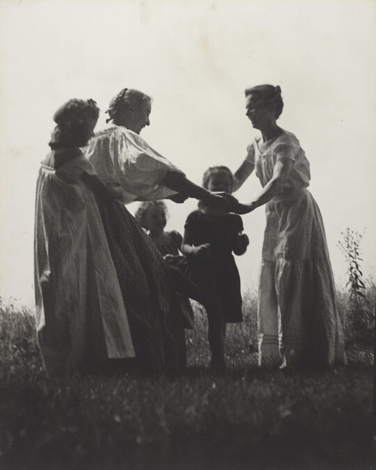 A black-and-white photograph of White women and children wearing long dresses holding hands to form a circle in a grassy field.