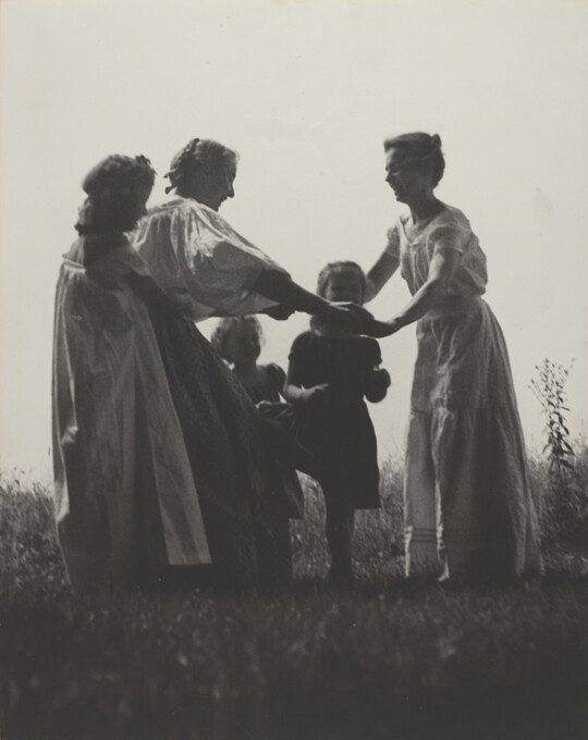 A black-and-white photograph of White women and children wearing long dresses holding hands to form a circle in a grassy field.