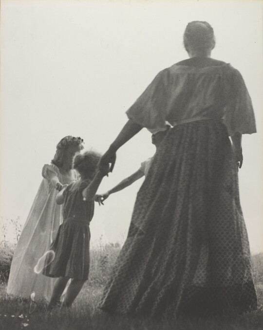 A black-and-white photograph of a woman, seen from the back, and two White children holding hands in a grassy field.