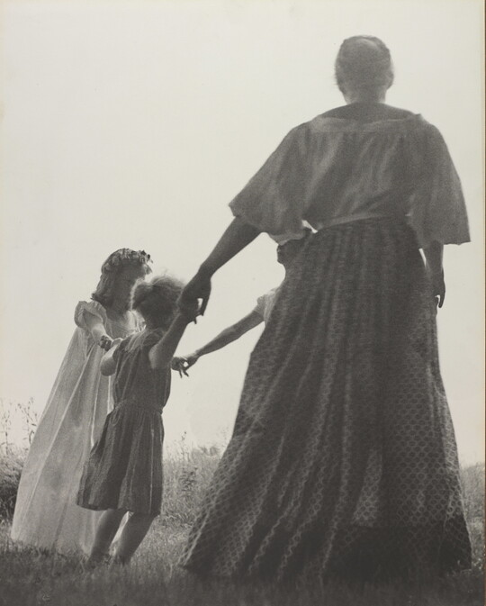 A black-and-white photograph of a woman, seen from the back, and two White children holding hands in a grassy field.