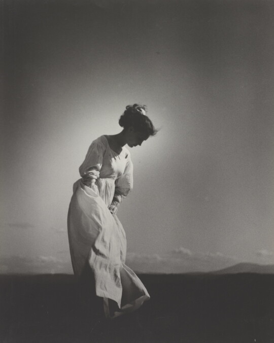 A black-and-white photograph of a White woman wearing a flowing white dress and looking down as she stands in a field.