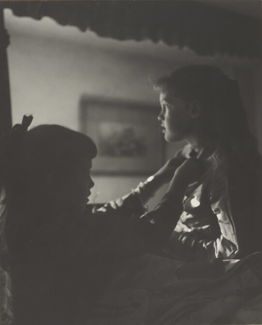 A black-and-white photograph of two young White girls, one fastening a ribbon around the neck of the other, both in shadow.