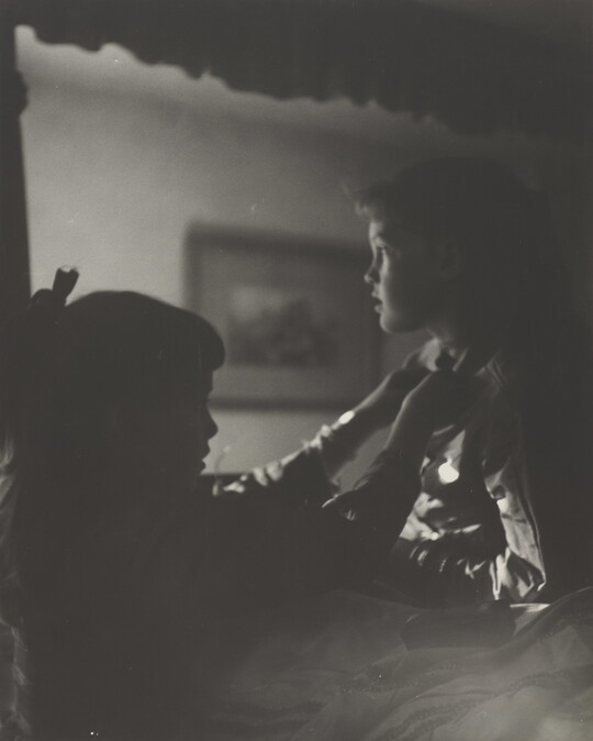 A black-and-white photograph of two young White girls, one fastening a ribbon around the neck of the other, both in shadow.