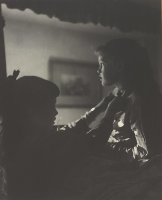 A black-and-white photograph of two young White girls, one fastening a ribbon around the neck of the other, both in shadow.