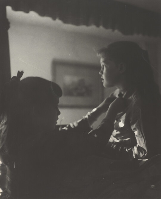 A black-and-white photograph of two young White girls, one fastening a ribbon around the neck of the other, both in shadow.