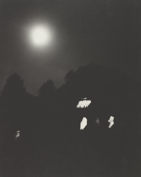 A black-and-white photograph of trees and a dark house with lit windows at night under a full moon.