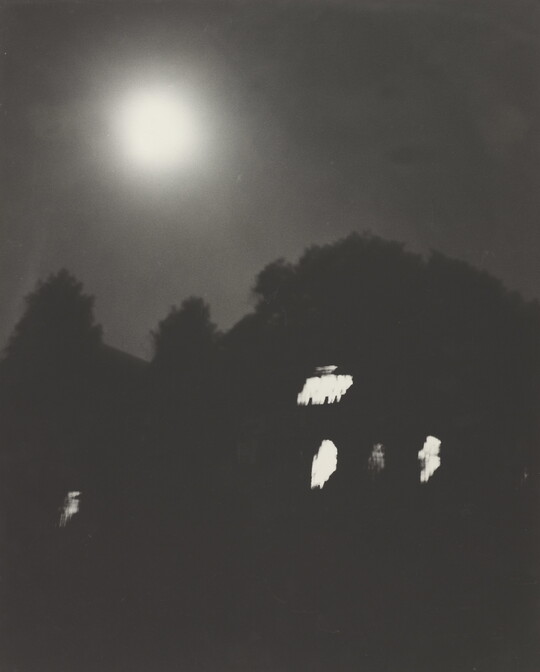 A black-and-white photograph of trees and a dark house with lit windows at night under a full moon.