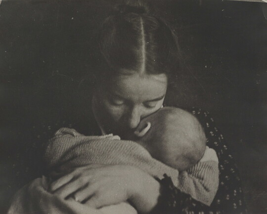A black-and-white photograph of a White woman holding an infant to her chest while she rests her cheek on the infant's cheek.
