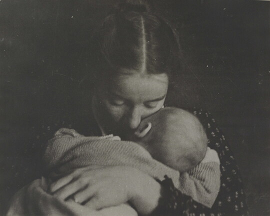 A black-and-white photograph of a White woman holding an infant to her chest while she rests her cheek on the infant's cheek.