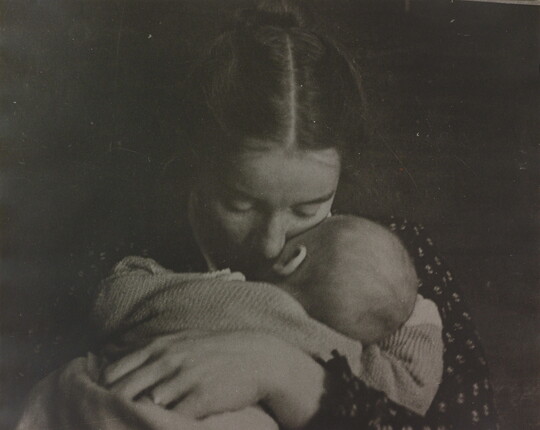 A black-and-white photograph of a White woman holding an infant to her chest while she rests her cheek on the infant's cheek.
