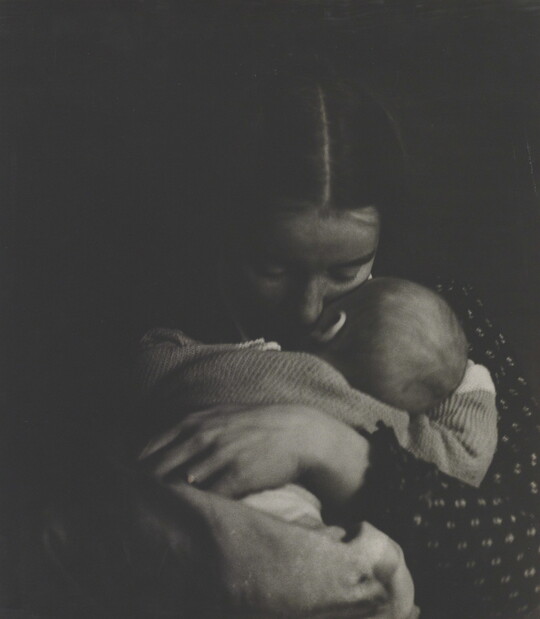 A black-and-white photograph of a White woman holding an infant to her chest while she rests her cheek on the infant's cheek.