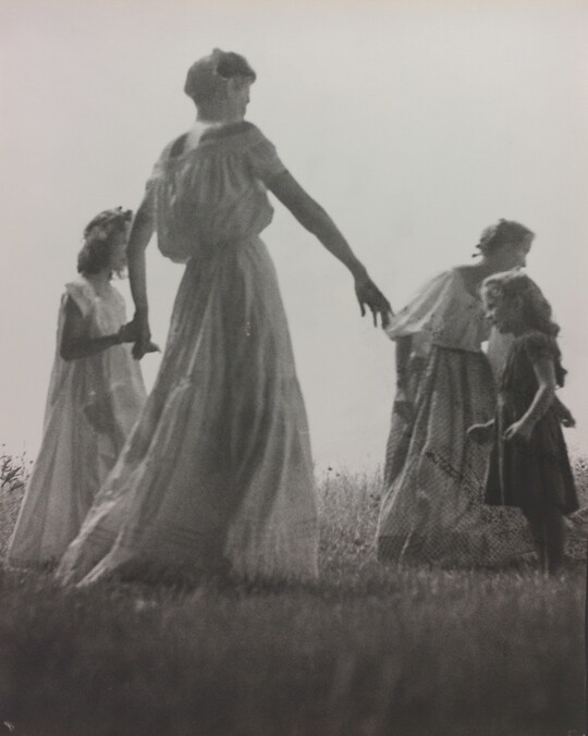 A black-and-white photograph of White women and children wearing long dresses holding hands to form a circle in a grassy field.