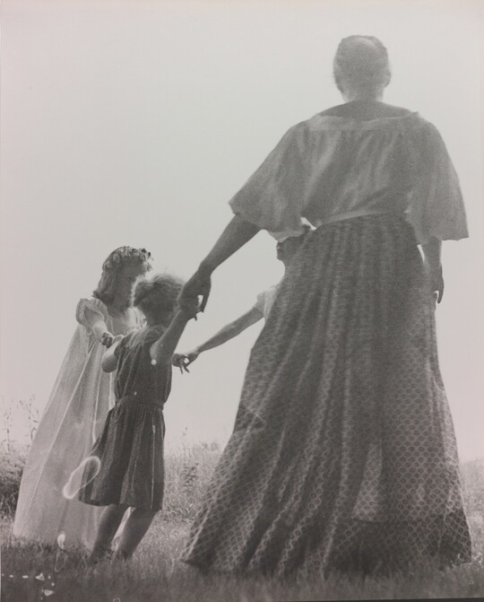 A black-and-white photograph of a woman, seen from the back, and two White children holding hands in a grassy field.