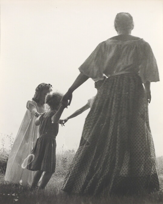 A black-and-white photograph of a woman, seen from the back, and two White children holding hands in a grassy field.