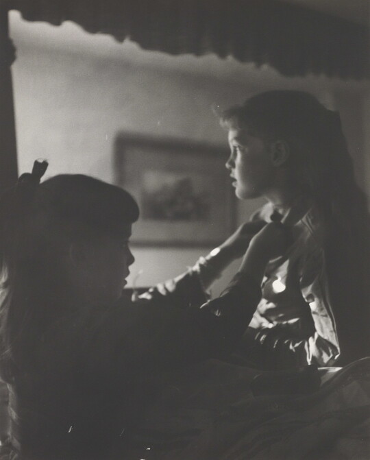 A black-and-white photograph of two young White girls, one fastening a ribbon around the neck of the other, both in shadow.