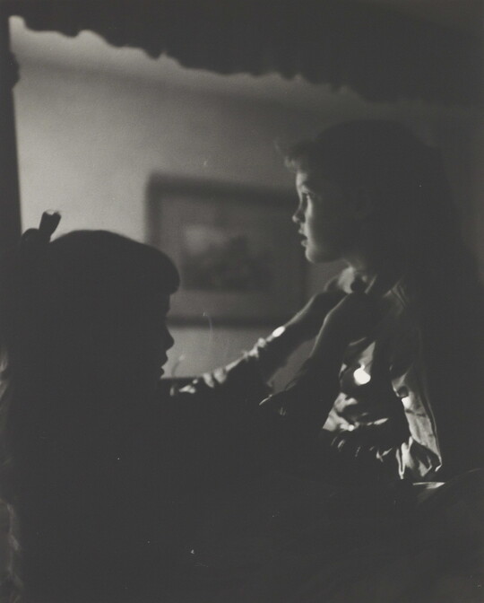 A black-and-white photograph of two young White girls, one fastening a ribbon around the neck of the other, both in shadow.