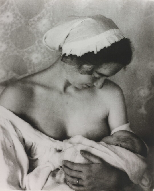 A black-and-white photograph of a White woman in a nightcap and white flowing blouse nursing an infant.