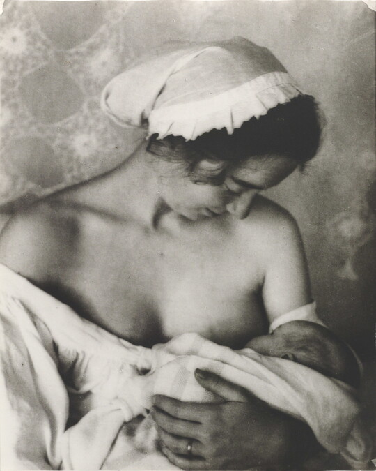 A black-and-white photograph of a White woman in a nightcap and white flowing blouse nursing an infant.