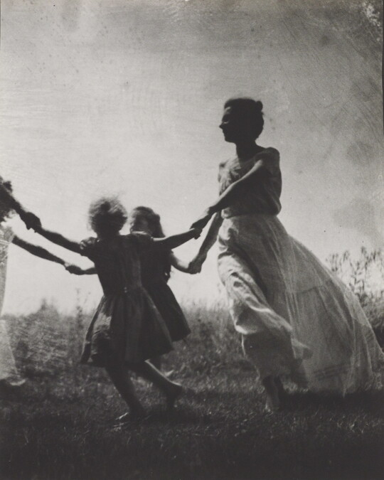 A black-and-white photograph of a White woman and children wearing long dresses holding hands to form a circle in a grassy field.