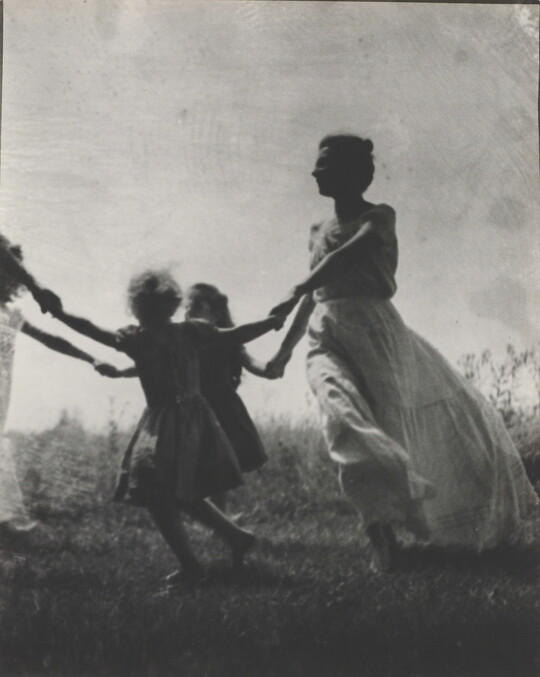A black-and-white photograph of a White woman and children wearing long dresses holding hands to form a circle in a grassy field.