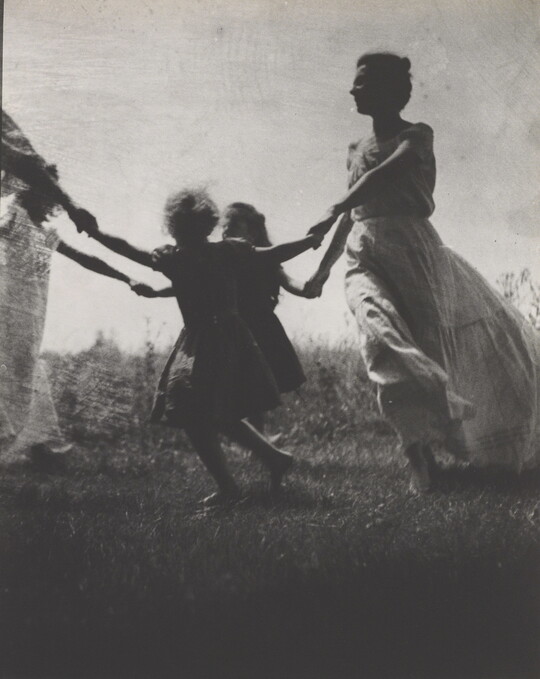 A black-and-white photograph of a White woman and children wearing long dresses holding hands to form a circle in a grassy field.