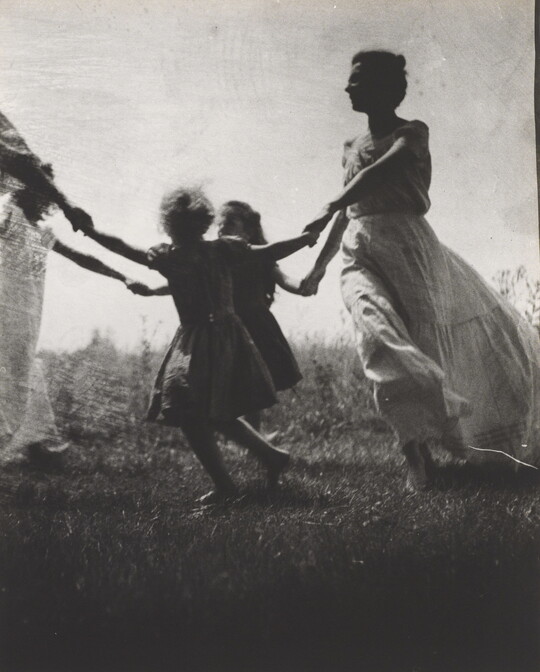 A black-and-white photograph of a White woman and children wearing long dresses holding hands to form a circle in a grassy field.