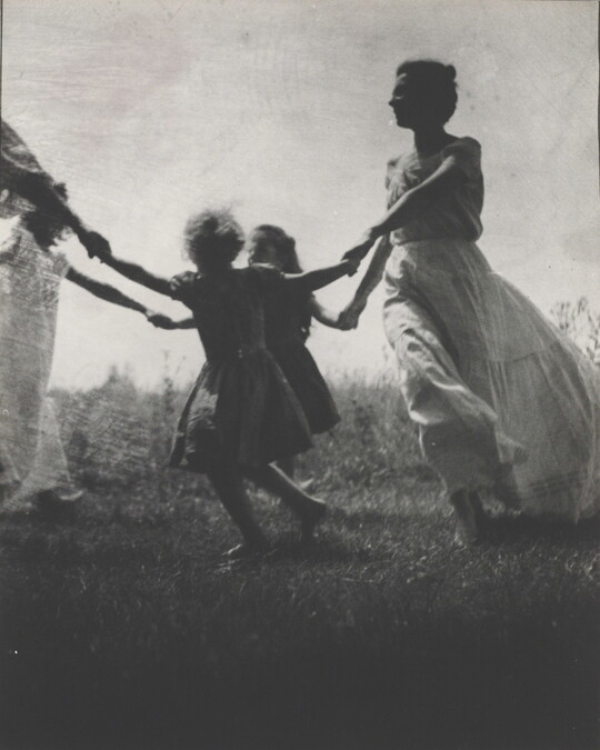 A black-and-white photograph of a White woman and children wearing long dresses holding hands to form a circle in a grassy field.