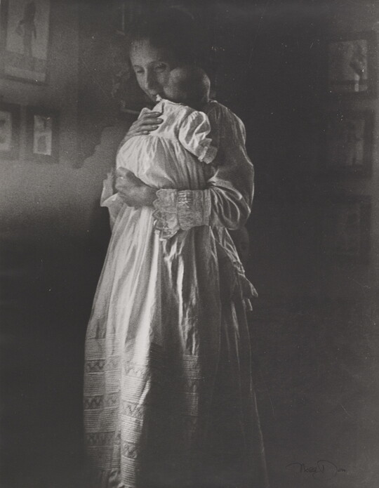 A black-and-white photograph of a standing White woman holding a baby wearing a long white nightgown.