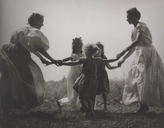 A black-and-white photograph of White women and children wearing long dresses holding hands to form a circle in a grassy field.