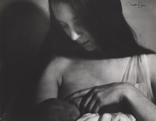 A black-and-white photograph of a White woman with long, dark hair breastfeeding a baby.