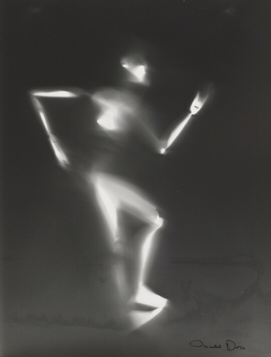 A black-and-white photograph of a white person-shaped puppet dancing on a black background.