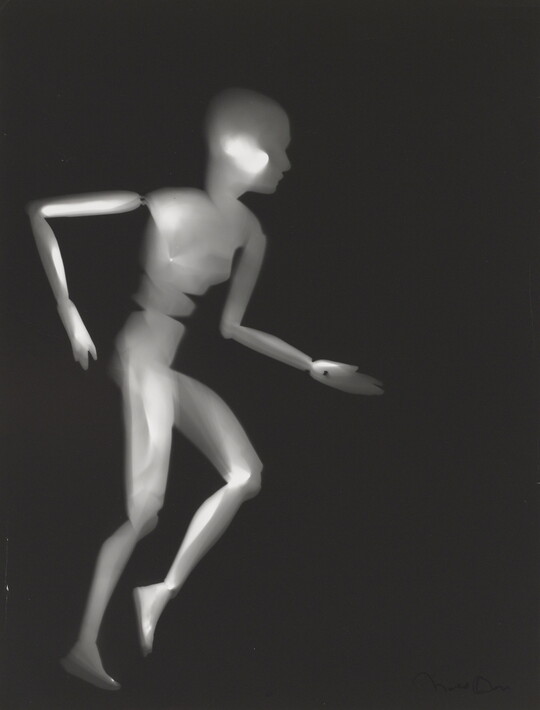 A black-and-white photograph of a white person-shaped puppet running on a black background.