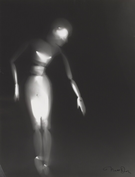 A black-and-white photograph of a white person-shaped puppet dancing on a black background.