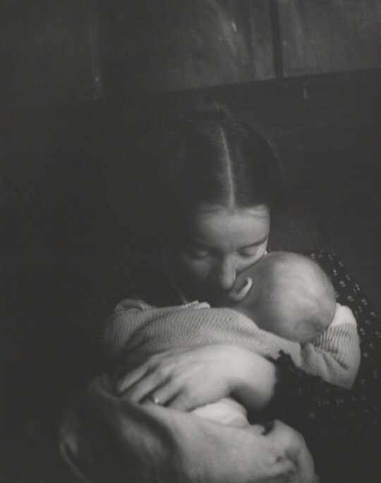 A black-and-white photograph of a White woman holding an infant to her chest while she rests her cheek on the infant's cheek.
