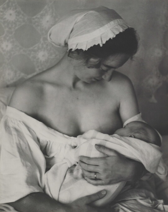 A black-and-white photograph of a White woman in a nightcap and white flowing blouse nursing an infant.