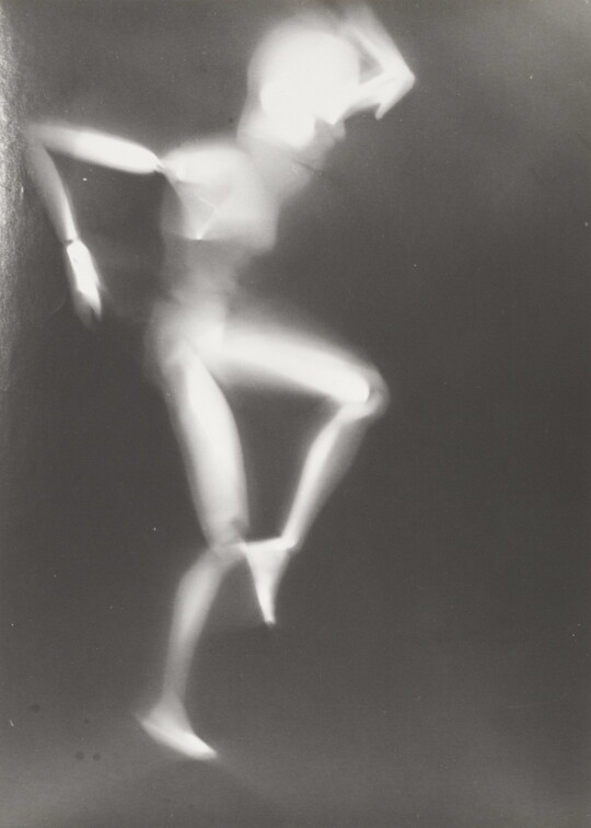 A black-and-white photograph of a white person-shaped puppet dancing on a black background.