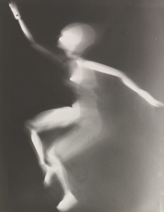 A black-and-white photograph of a white person-shaped puppet dancing on a black background.