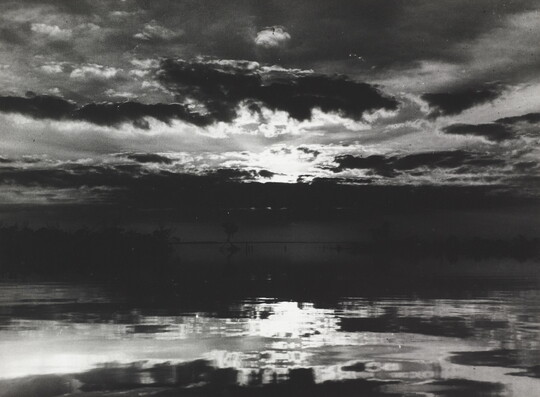 A black-and-white photograph of a cloudy sky with sun peeking through reflected on water below.