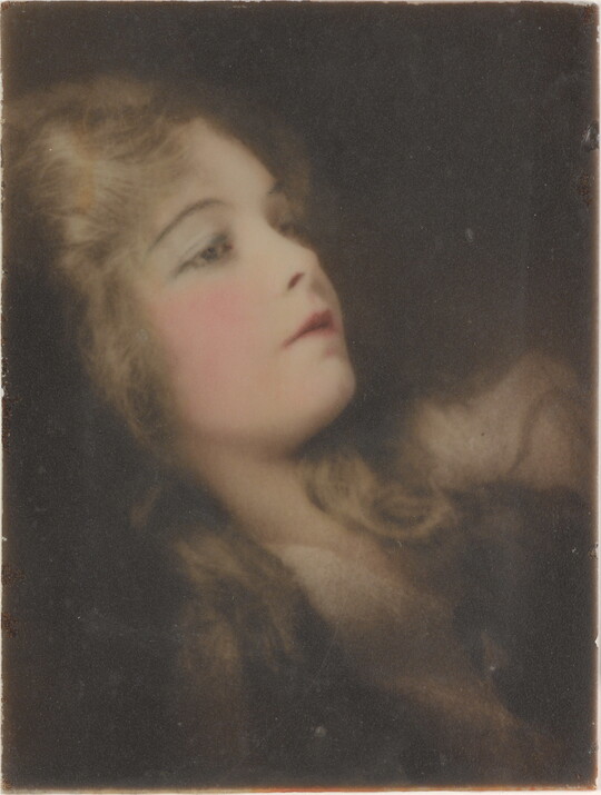 A hand-colored, cropped photograph of a reclining woman with long hair and flushed pink cheeks.