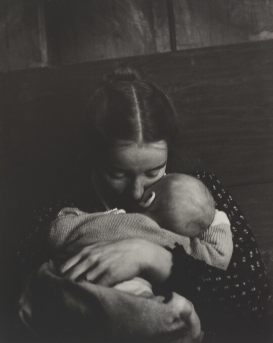 A black-and-white photograph of a White woman holding an infant to her chest while she rests her cheek on the infant's cheek.