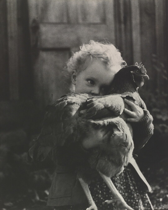 A black-and-white photograph of a White toddler holding a chicken close and obscuring part of her face.