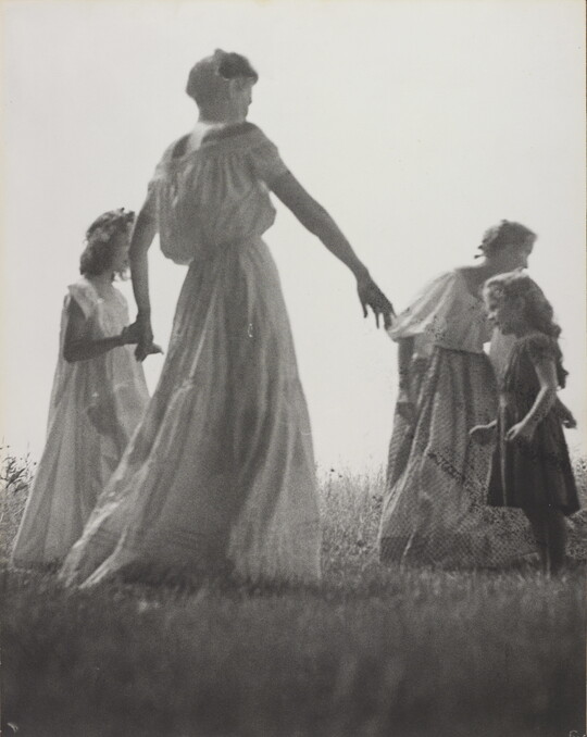 A black-and-white photograph of White women and children wearing long dresses holding hands to form a circle in a grassy field.