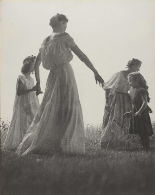 A black-and-white photograph of White women and children wearing long dresses holding hands to form a circle in a grassy field.