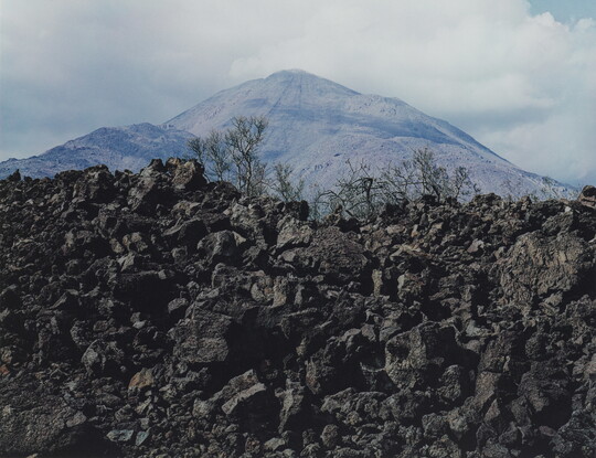 A color photograph of a hill of black stones, the tops of some evergreens, and a mountain peak in the background.