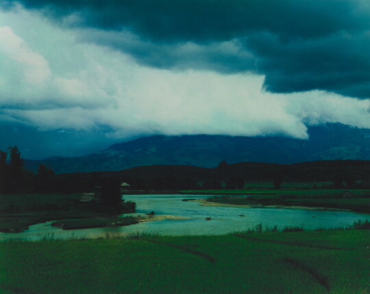A color photograph of storm clouds rolling over a mountain with a winding river in the foreground.