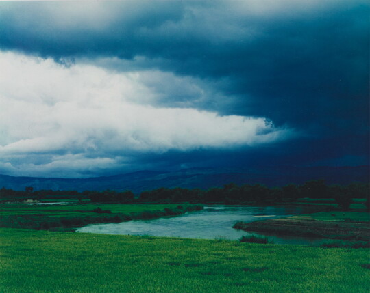 A color photograph of storm clouds rolling over a mountain with a winding river in the foreground.