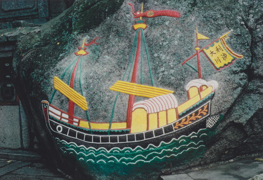 A color photograph of a boat with three red masts, yellow and red flags sailing on wavy green water all painted onto a rock.