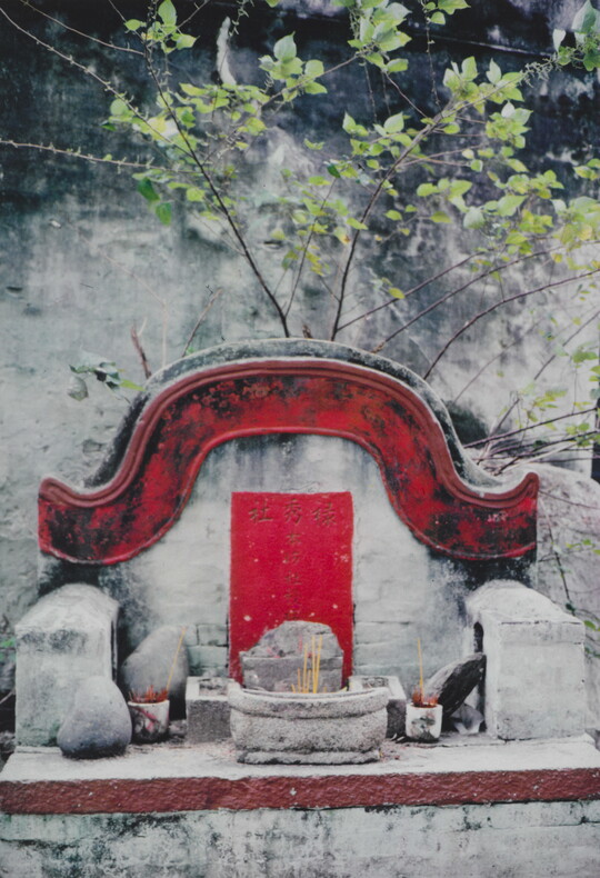 A color photograph of a stone altar with a red border above it and several incense sticks in various small vessels.