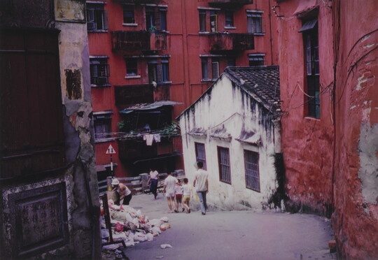A color photograph of people walking down a garbage-strewn sloping street in front of tall red buildings and a single-story white building.